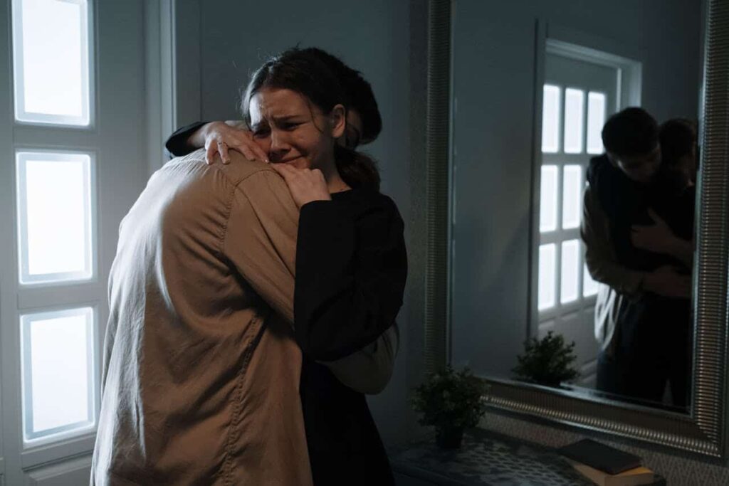Two people embrace tightly in a dimly lit room by a window, both appearing emotional. One person is crying from overwhelming anxiety triggers as they hold the other, their reflection visible in a large mirror nearby.